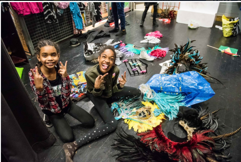 two black girls under the age of twelve sitting on the floor backstage smiling with their colourful costumes on the floor
